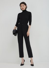 Top noir ajusté à col roulé avec manches longues, associé à un pantalon taille haute sur mesure noir. Le mannequin tient une pochette noire et porte des talons noirs.