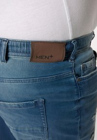Jeans de mezclilla con un lavado azul claro, que cuentan con una parche de cuero marrón etiquetado "MEN+" en la cinturilla y un detalle en el bolsillo trasero.