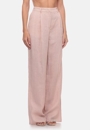 Donna che indossa pantaloni rosa chiaro a vita alta, a gamba larga con pieghe anteriori e sandali aperti davanti, in piedi su uno sfondo semplice.