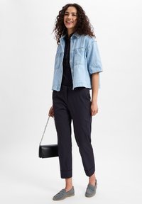 Helles blaue Jeansjacke im cropped Stil, schwarzes Poloshirt, marineblaue Hose mit hochgekrempelten Beinen, graue Loafer und eine schwarze Handtasche mit Kettenriemen.