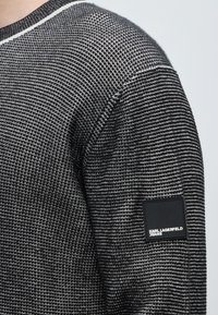 Manche de pull en tricot texturé noir et blanc avec un patch rectangulaire noir portant l'inscription Karl Lagerfeld Jeans sur le haut du bras.