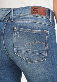 Närbild av bakfickan på blå denimjeans med lätt slitage och en brun läderpatch med texten "RAW" på midjebandet.