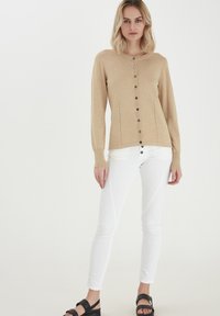 Beige kardigan s kulatým výstřihem, zapínáním na knoflíky vpředu a detailním bočním švem, spárován s bílými úzkými džíny a černými sandály.