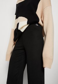 Schwarze gerippte Hose mit tailliertem Design, kombiniert mit einem farblich abgestimmten Strickpullover in Beige, Schwarz und Creme. Mit Logodetail.