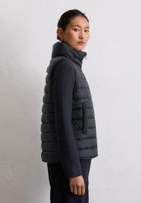 Femme de profil portant un gilet sans manches noir matelassé par-dessus un haut à manches longues noir, sur un fond mur léger et uni.