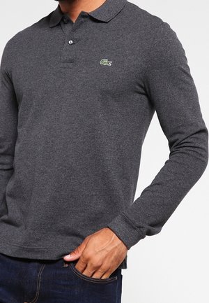 Mężczyzna ubrany w ciemnoszarą polówkę z długim rękawem z zielonym logo krokodyla na piersi, ręka spoczywa w kieszeni niebieskich dżinsów.