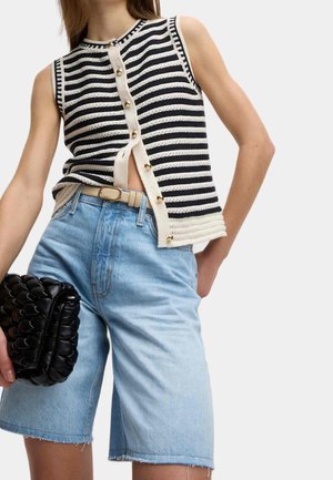 Vrouw met een mouwloos, zwart-wit gestreept, geknoopt topje, lichtblauwe high-waisted denim shorts, beige riem, en een zwarte geprofileerde clutch in haar hand.