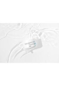 Crème enrichie en puissance d'hydratation dans un pot rond blanc, avec du texte noir et une accentuation bleue, flottant dans de l'eau claire avec de légères ondulations.