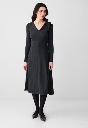 Kobieta ubrana w ciemnoszarą, długorękawową sukienkę midi z dekoltem w kształcie litery V i kwiatowym zdobieniem na lewym ramieniu, czarne rajstopy oraz metaliczne, szpiczaste buty.