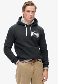 Szary polar z kapturem i sznurkami, z przednią kieszenią oraz białym logo "Vintage SuperDry". Nałożono na niego szary T-shirt, zestawiony z beżowymi spodniami.