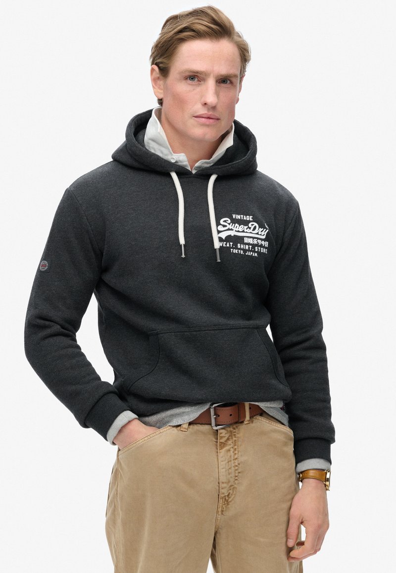 Szary polar z kapturem i sznurkami, z przednią kieszenią oraz białym logo "Vintage SuperDry". Nałożono na niego szary T-shirt, zestawiony z beżowymi spodniami.