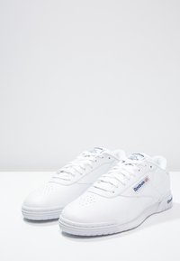 Baskets en cuir blanc avec embout perforé, lacets plats et texture lisse. Présentent des accents bleus et un logo sur le côté. Design classique.