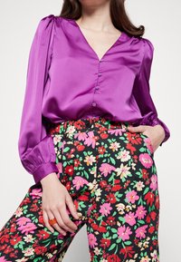 Blusa di raso viola con scollatura a V e chiusura davanti con bottoni; abbinata a pantaloni floreali neri in rosso, rosa e verde con tasche laterali.