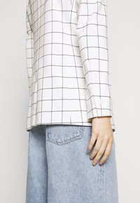 Chemise blanche à manches longues avec motif à carreaux noirs, dotée de poignets à boutons. Associée à un short en denim bleu clair avec des détails de poche arrière.