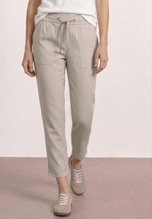 Kobieta nosząca jasnobeżowe spodnie z troczkiem i przednimi naszywanymi kieszeniami oraz jasnobeżowe trampki, idąca po gładkiej podłodze.