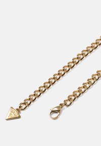 Gouden schakelarmband met een karabijnsluiting en een kleine driehoekige hanger met de gravure "GUESS" en een vraagteken.