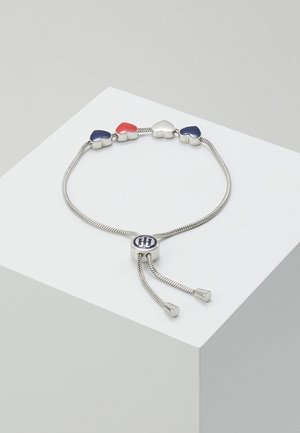 Sølvjusterbart armbånd med fire hjerteformede vedhæng i rød, hvid og blå, med en rund lukning med en blå logo.