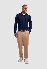 Polo a maniche lunghe lavorata a maglia blu navy con motivo a rombi, pantaloni beige plissettati e sneakers bianche con dettagli neri. Una cintura semplice completa il look.