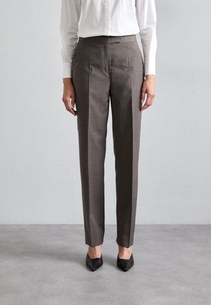 Persona que lleva puestos pantalones de vestir marrones a medida, camisa blanca de manga larga y zapatos negros de punta afilada, de pie sobre un suelo gris frente a una pared lisa.
