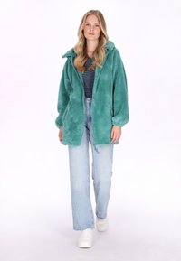 Chaqueta de forro polar esponjosa en color verde azulado con capucha y cierre de cremallera. Llevada sobre una camiseta de rayas y combinada con vaqueros azul claro y zapatillas blancas.