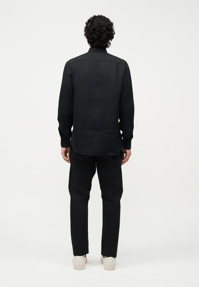 Άνδρας με σγουρά μαλλιά, ντυμένος με μαύρο μακρυμάνικο πουκάμισο, μαύρο παντελόνι και λευκά αθλητικά παπούτσια, στέκεται με την πλάτη προς το μέρος μας μπροστά σε απλό λευκό φόντο.