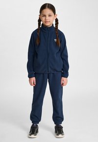 Marinblå fleecedräkt i två delar som inkluderar en jacka med dragkedja och hög krage samt matchande byxor med elastiska manschetter. Sneakers fulländar stilen.