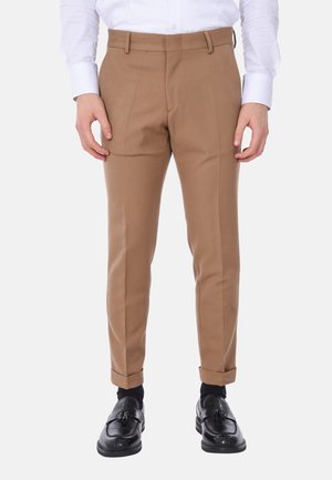Pantaloni beige dalla vestibilità slim, con pieghe frontali e risvolti. Abbinati a mocassini neri, indossati sopra calzini scuri.