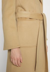 Beige Wollmantel mit tailliertem Schnitt, weiten Ärmeln und seitlichen Taschen, ausgestattet mit einer sanften Textur und einer strukturierten Silhouette.