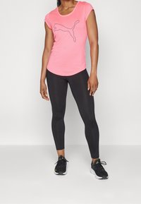 Kvinna klädd i en rosa Puma-t-shirt, svarta leggings och svarta sportskor som står mot en vit bakgrund.