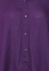 Lila skjorta med knappslå och en slät yta, med sex ljusa knappar som löper vertikalt ner längs framsidan.