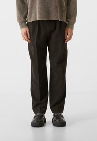 Pantalones anchos de tiro alto en negro con pliegues marcados, hechos de una tela suave. Combinados con zapatos negros gruesos para un look moderno.