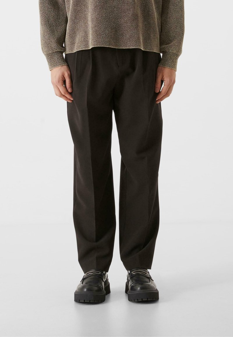 Pantalones anchos de tiro alto en negro con pliegues marcados, hechos de una tela suave. Combinados con zapatos negros gruesos para un look moderno.