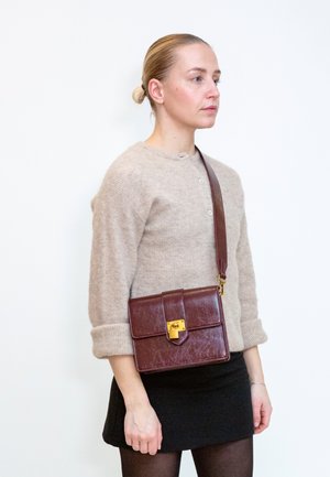 Femme aux cheveux blonds en chignon porte un pull beige et une jupe noire, elle porte un sac bandoulière en cuir marron foncé avec une serrure dorée.