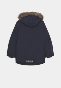 Manteau bleu marine avec une capuche doublée en fourrure, manches longues et un logo au bas. Le matériau semble lisse avec une finition mate.