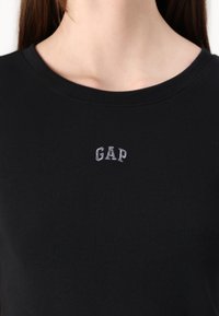 T-shirt noir à côtes avec un col rond et un petit logo "GAP" brodé en gris sur la poitrine. Texture lisse, coupe décontractée.