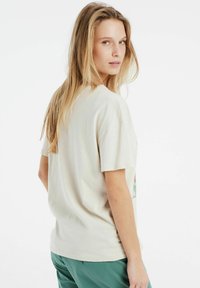 Camiseta de algodón color beige claro con mangas cortas, corte relajado y un diseño impreso que presenta acentos verdes en la parte delantera.