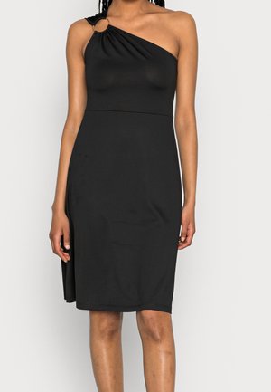 Femme portant une robe noire d'une épaule, longueur genou, avec un détail d'anneau sur la bretelle, debout devant un fond clair uni.