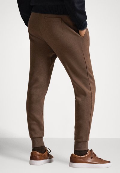 Brązowe, zwężane spodnie dresowe z bocznymi kieszeniami i ściągaczami u dołu, wykonane z miękkiego materiału. Połączone z jasnobrązowymi tenisówkami i ciemnym swetrem.