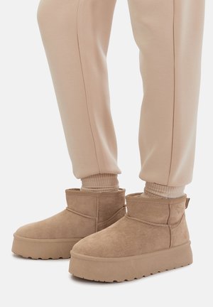 Braune Wildleder-Stiefeletten mit strukturierter Oberfläche und dicker Plateau-Sohle. Kombiniert mit beigen Jogginghosen und passenden Knöchelsocken.