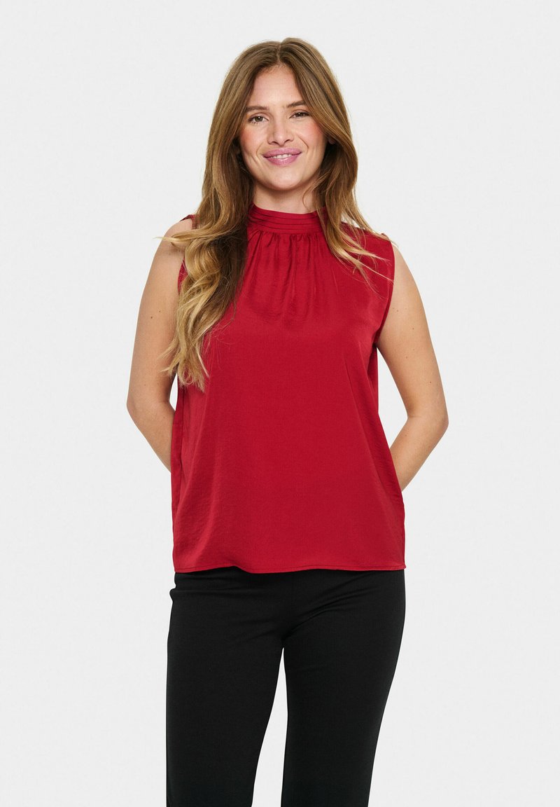 Top rosso senza maniche con un colletto alto e dettagli arricciati, abbinato a pantaloni neri. Il tessuto appare liscio e leggero.