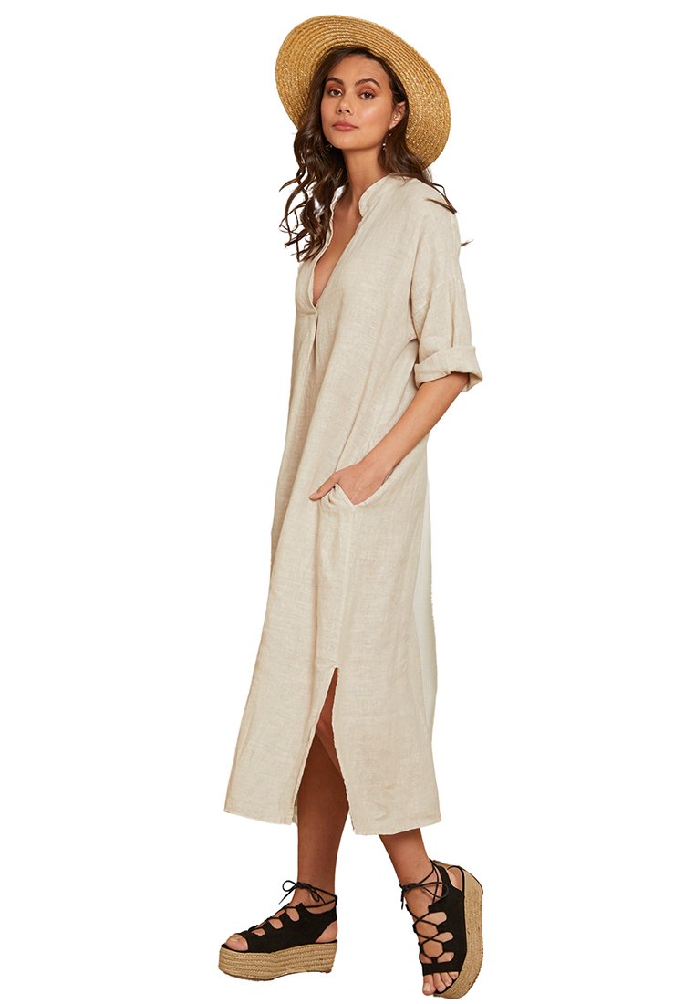 Beiges Leinenkleid mit V-Ausschnitt, gerafften Ärmeln, seitlichen Taschen und seitlichen Schlitzen. Kombiniert mit schwarzen Keilsandalen und einem Strohhut.