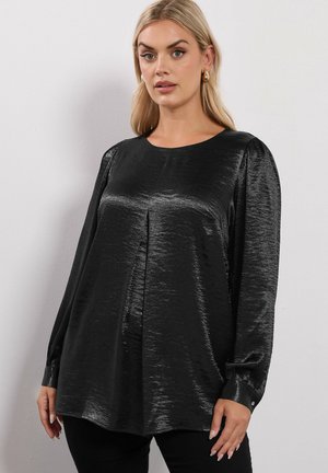 Blouse noire à manches longues avec une texture brillante, un col rond et des détails de plis. Présente des accents de boutons sur les poignets.
