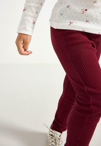 Burgundin sävyiset ribattua corduroita olevat housut, joissa on tiukka istuvuus, yhdistettynä vaaleisiin nauhallisiin tennareihin. Valkoinen kukkakuosinen pitkähihaisuus toppi näkyy osittain.