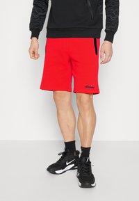 Hombre que lleva puestos pantalones cortos rojos brillantes con el logo de ellesse, calcetas negras, zapatillas Nike negras y un sudadera negra con mangas estampadas.