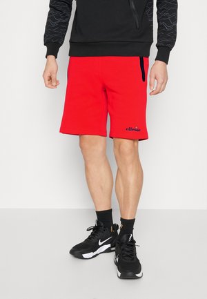 Hombre que lleva puestos pantalones cortos rojos brillantes con el logo de ellesse, calcetas negras, zapatillas Nike negras y un sudadera negra con mangas estampadas.