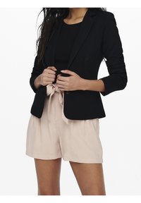 Blazer nero aderente con rever a punta, abbinato a un top nero aderente e pantaloni corti a vita alta rosa chiaro con dettaglio della cintura legata.