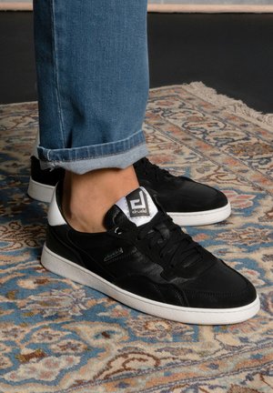 Person wearing black and white sneakers with logo on tongue, standing on a patterned rug with blue rolled-up jeans.