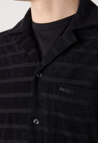 Gros plan d'une personne portant une chemise noire à col avec de subtiles rayures horizontales et une poche poitrine brodée de "HUGO" sur le côté gauche.