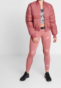 Chaqueta bomber rosa acolchada con puños acanalados, combinada con leggings rosas con detalles brillantes y un top deportivo negro. Zapatillas deportivas negras.