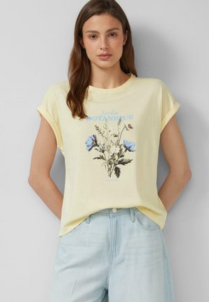 Kobieta o długich brązowych włosach, ubrana w bladożółtą koszulkę z niebieskim kwiatowym nadrukiem i jasnoniebieskie dżinsy, stoi na jednolitym tle.